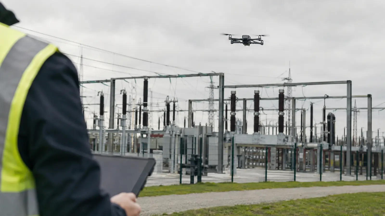 Inspektionsdrohne im Schwebeflug vor einer Umspannanlage, im Vordergrund unscharf eine Person mit Warnweste und Tablet.