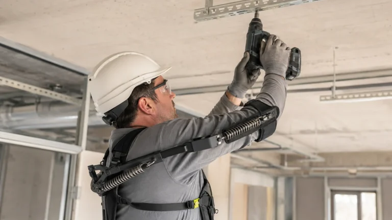 Bauarbeiter mit generischem passivem Überkopf-Exoskelett bohrt über Kopf auf einer Baustelle.